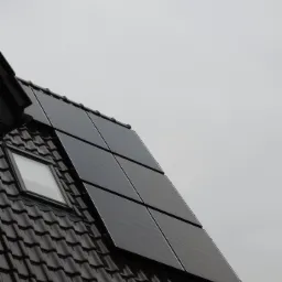 Ciemne panele słoneczne na brązowym dachu z dachówki ceramicznej, obok okno dachowe, pochmurne niebo w tle.