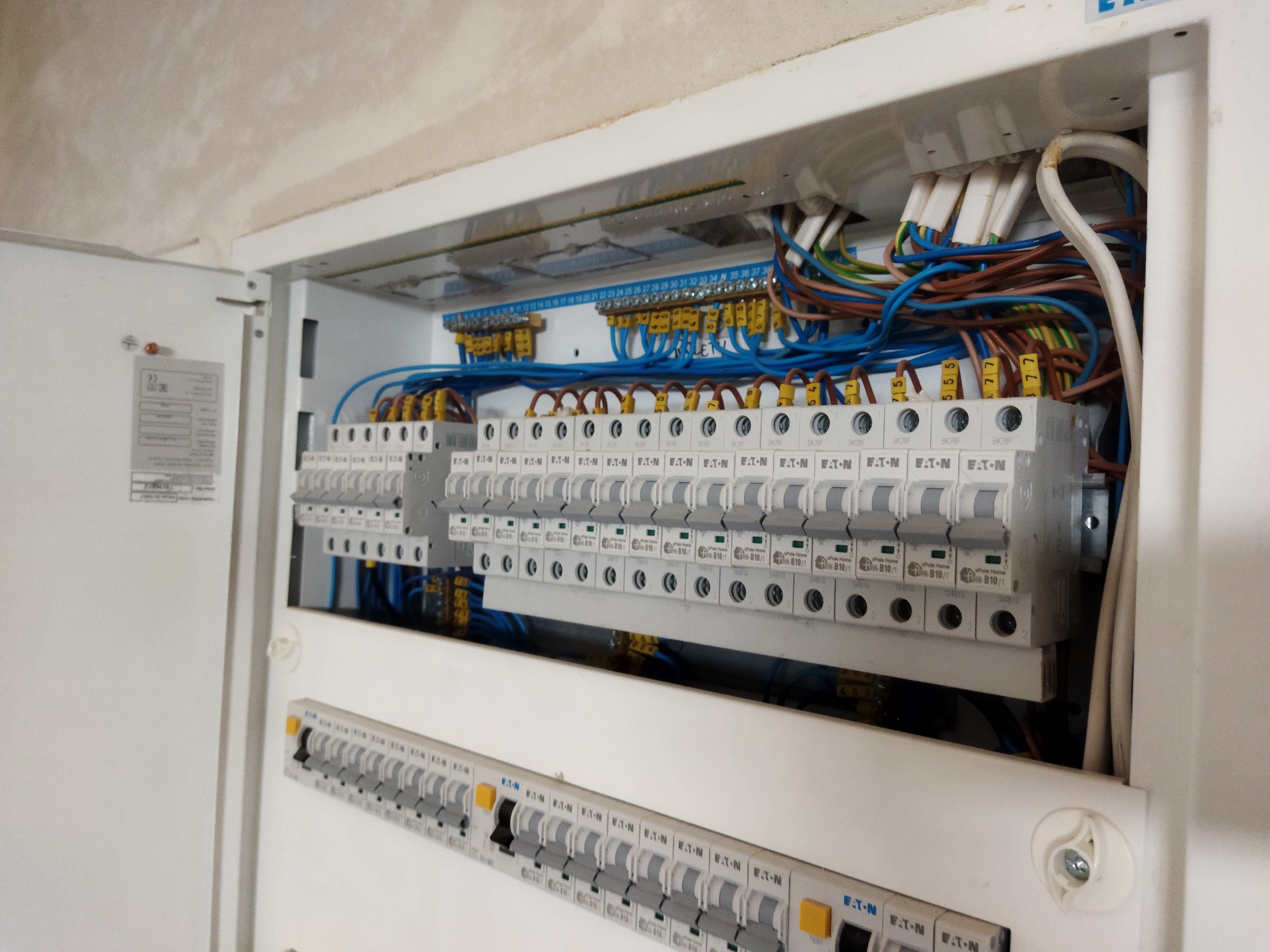 Otwarta skrzynka rozdzielcza z licznymi bezpiecznikami i okablowaniem, widoczne przewody w różnych kolorach oraz listwy zaciskowe, zamontowana w jasnym pomieszczeniu.