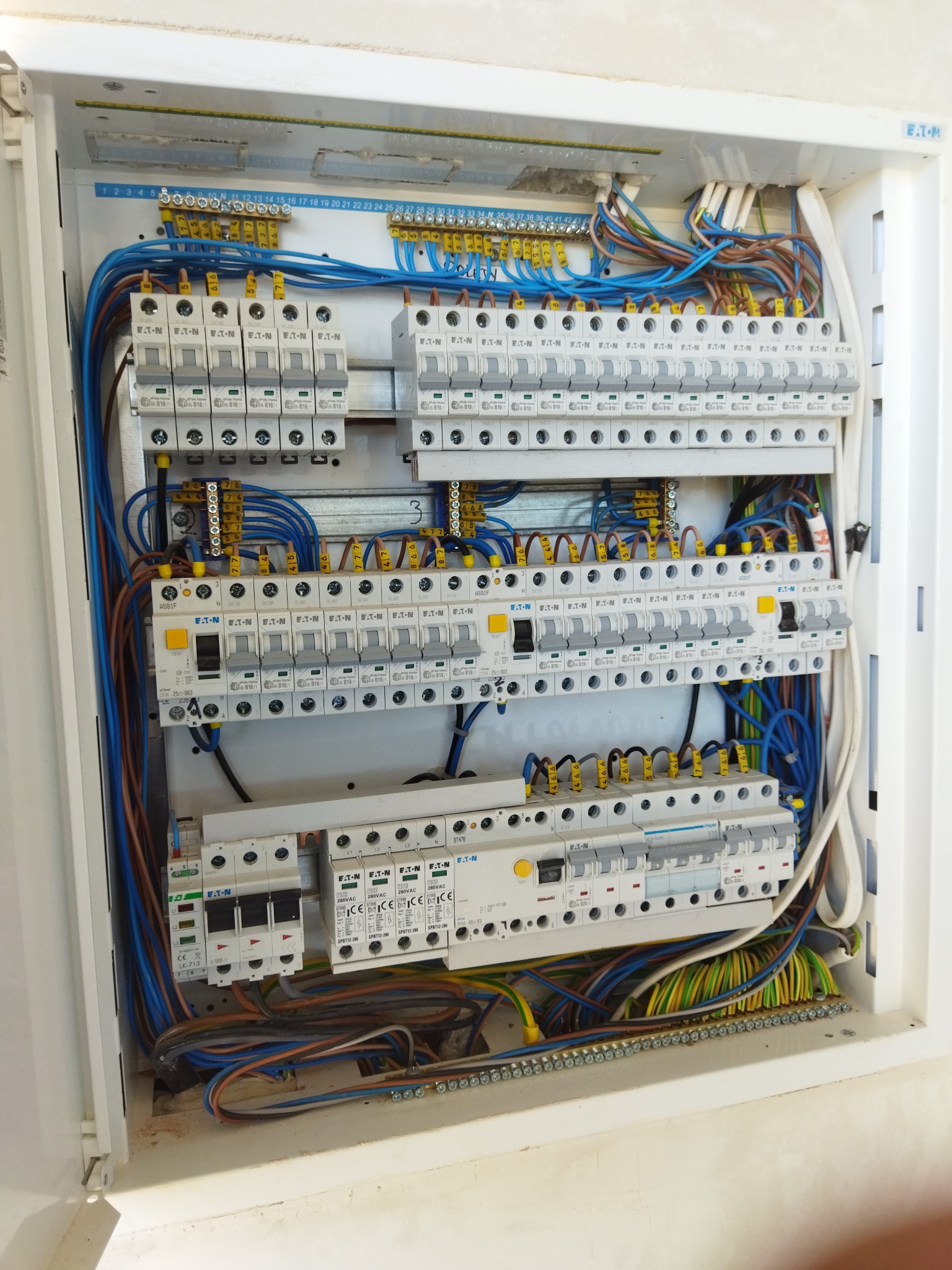 Otwarta skrzynka z rozbudowaną instalacją elektryczną, widoczne liczne zabezpieczenia i okablowanie w kolorach niebieskim, brązowym i zielono-żółtym, przewody uporządkowane i podłączone do listew...