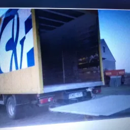 Żółty samochód dostawczy z otwartą rampą załadunkową i niebieskim logo firmy na boku, zaparkowany na trawiastym terenie z budynkiem w tle.