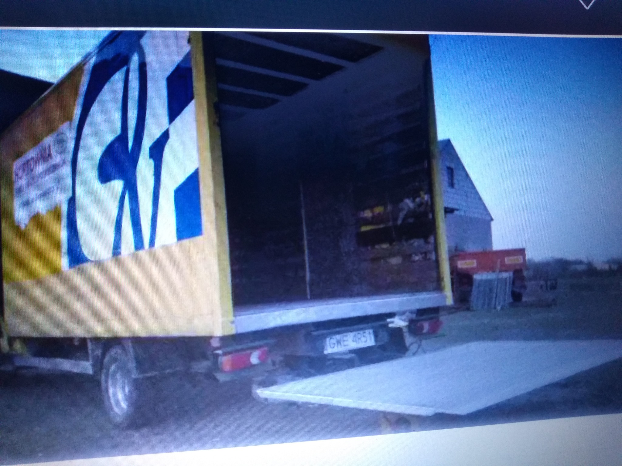 Żółty samochód dostawczy z otwartą rampą załadunkową i niebieskim logo firmy na boku, zaparkowany na trawiastym terenie z budynkiem w tle.