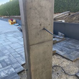 Piotr Orzechowski - Betonowy słupek ogrodzeniowy z widocznymi przewodami, częściowo ułożona kostka brukowa i zielony żywopłot w tle. Prace w toku, widoczne narzędzia i materiały budowlane.