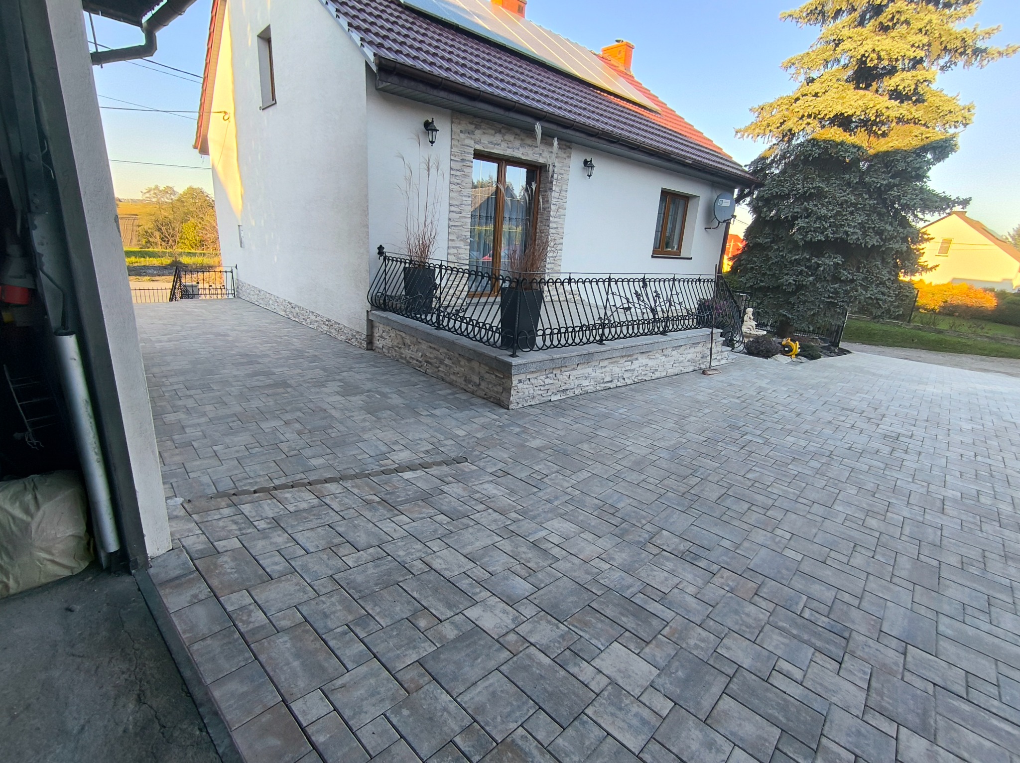 Ułożona szara kostka brukowa na podjeździe przed domem z balkonem i dekoracyjnym ogrodzeniem, w tle drzewo iglaste i błękitne niebo.