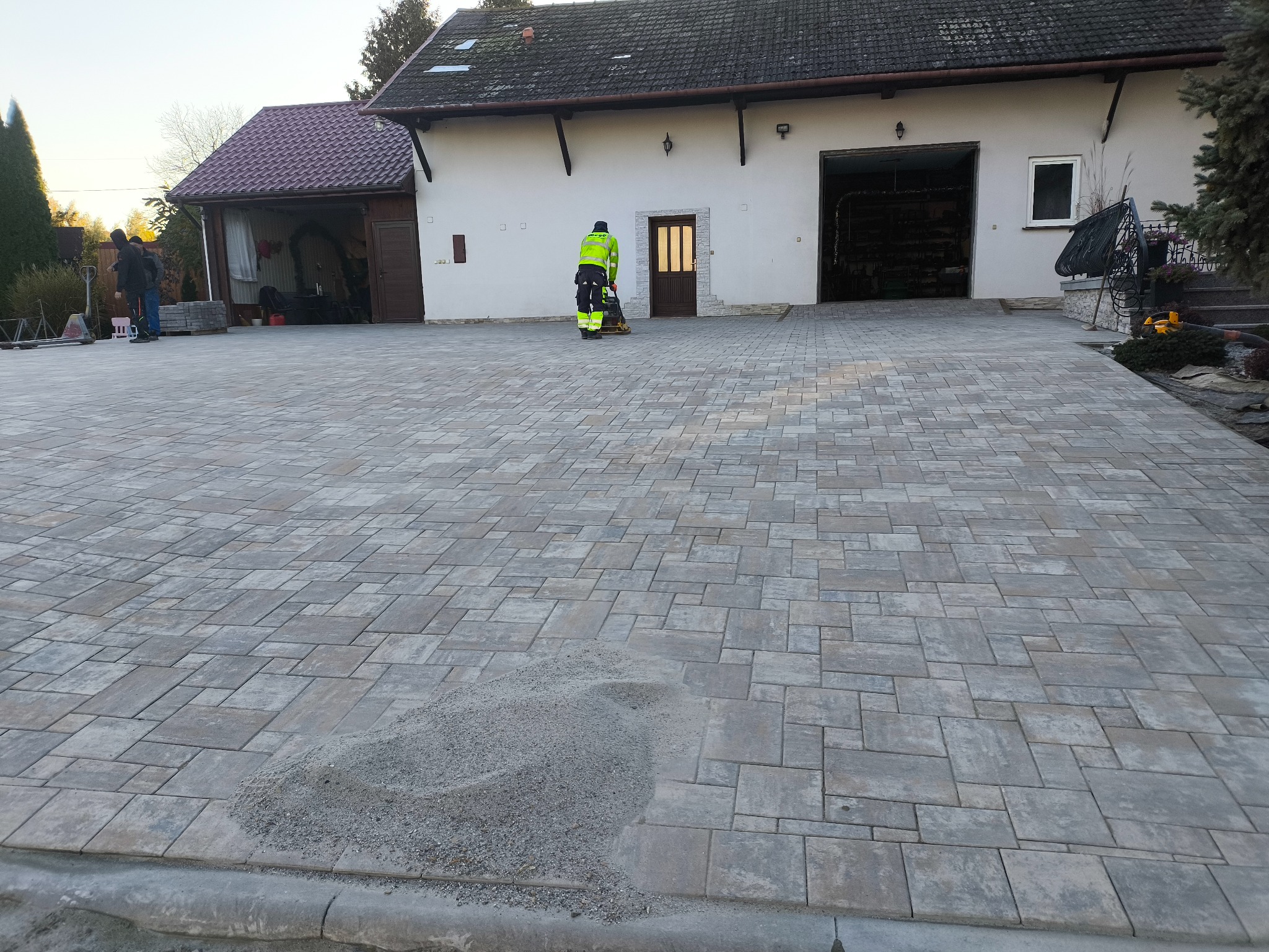 Prace brukarskie: zagęszczarka w użyciu na świeżo ułożonej kostce brukowej przed budynkiem z garażem. Widoczny stos piasku i pracownicy w tle.