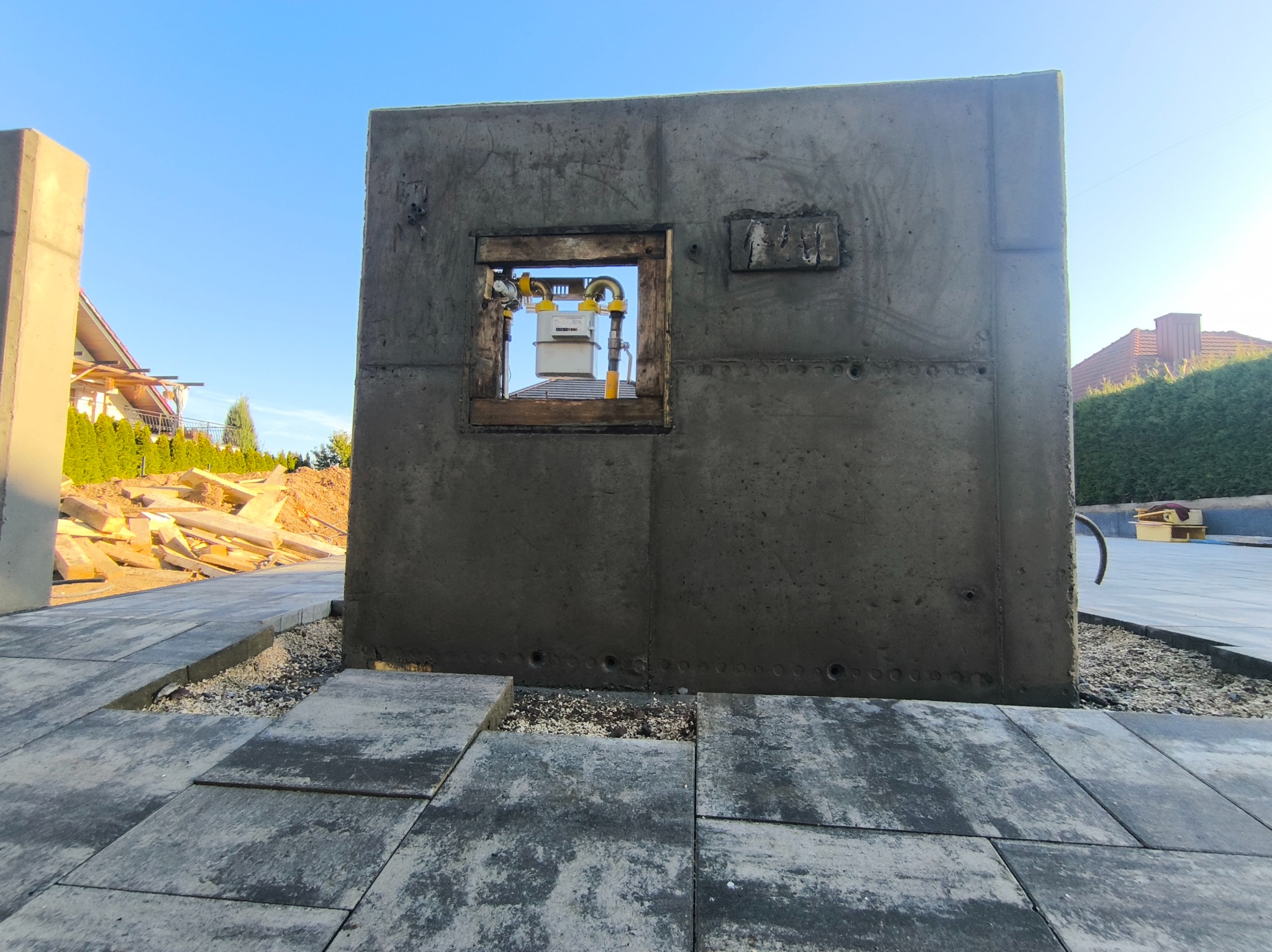 Betonowy mur z okienkiem na gazomierz w trakcie układania kostki brukowej. Widoczne elementy instalacji gazowej i fragmenty domu w tle.