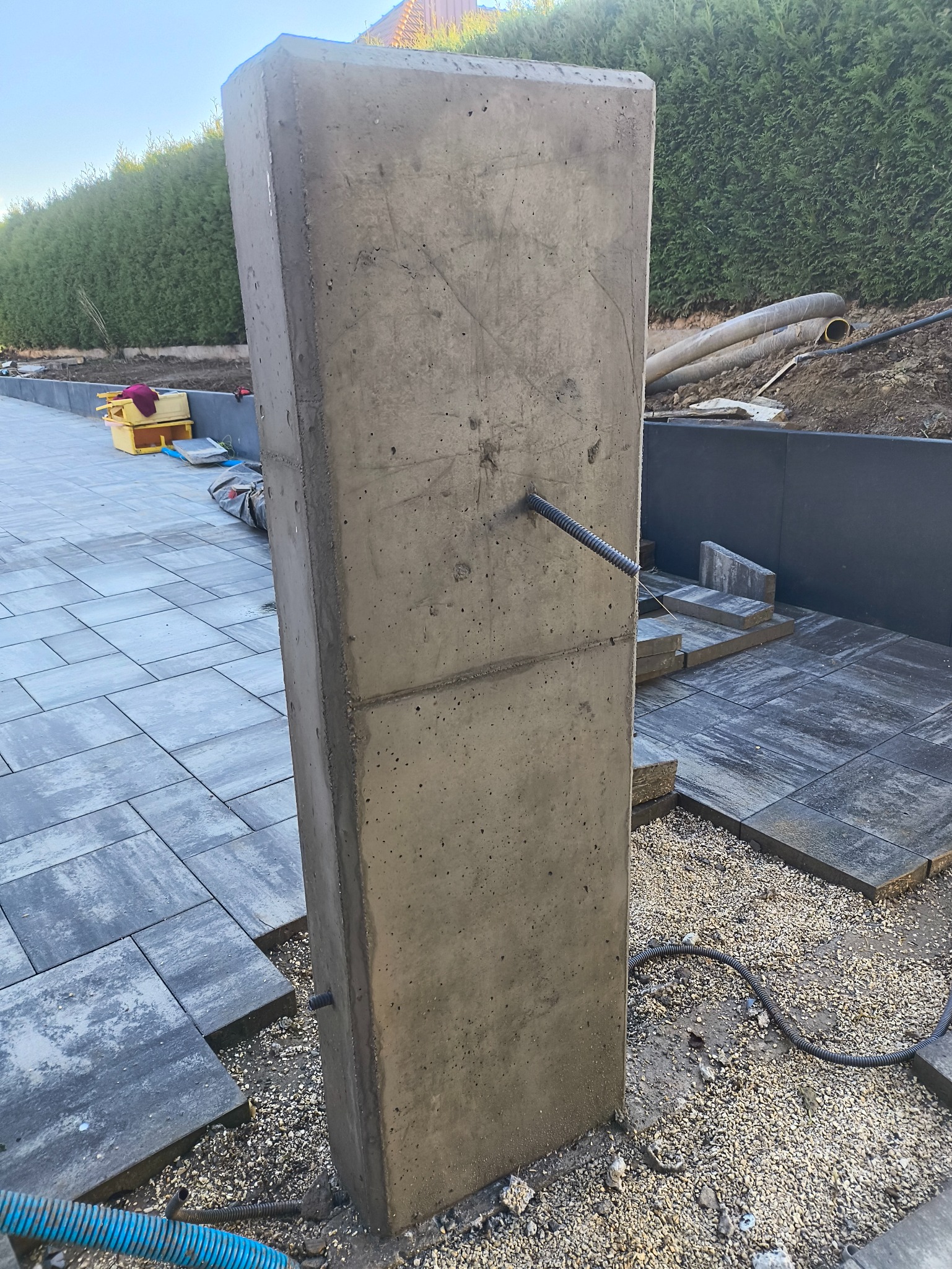 Betonowy słupek ogrodzeniowy z widocznymi przewodami, częściowo ułożona kostka brukowa i zielony żywopłot w tle. Prace w toku, widoczne narzędzia i materiały budowlane.