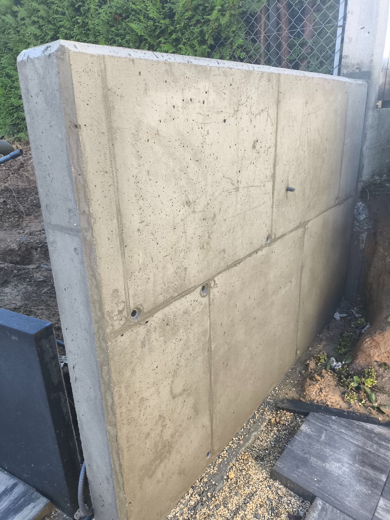 Betonowy murek oporowy z widocznymi otworami drenażowymi, oddzielający teren, z siatką ogrodzeniową w tle i fragmentami kostki brukowej u podstawy.