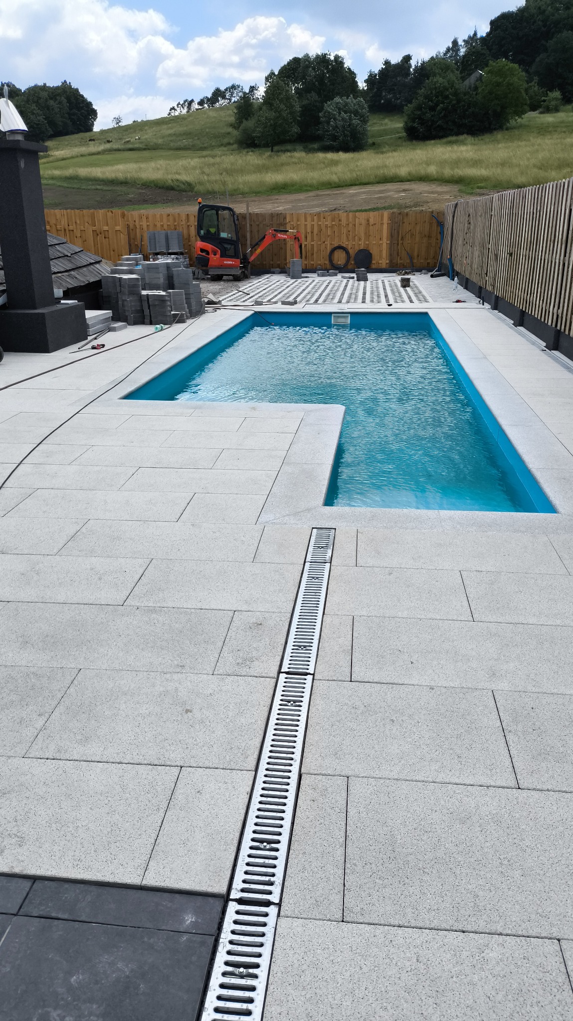 Nowoczesny basen z jasnoszarą kostką brukową i odpływem liniowym. W tle widoczny mini-koparka i paleta z kostką, co sugeruje trwające prace wykończeniowe.