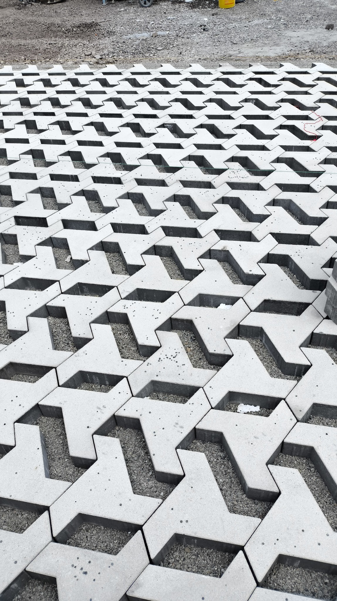 Ułożona, betonowa kostka ażurowa z wypełnieniem żwirowym. Widoczny fragment placu budowy z piaskiem i sprzętem. Nietypowy wzór kostki, powtarzalny układ.