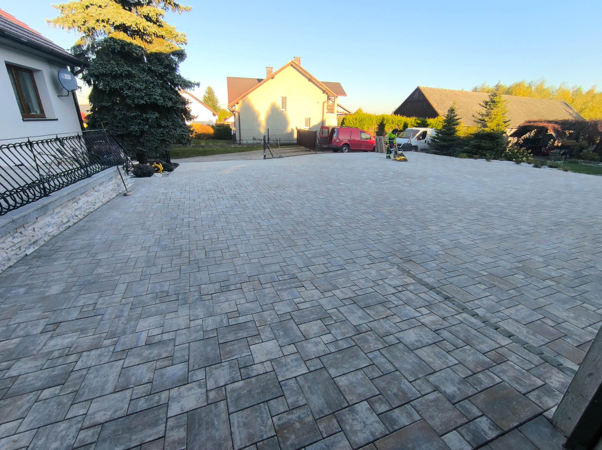 Rozległy podjazd z szarej kostki brukowej ułożonej we wzór, z widocznymi budynkami w tle i pracownikiem używającym zagęszczarki. Całość w słoneczny dzień.