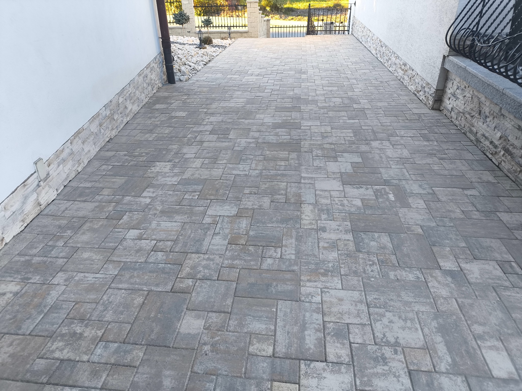 Ułożona kostka brukowa w odcieniach szarości tworząca wzór na podjeździe. Widoczne obrzeża z kamienia dekoracyjnego i brama wjazdowa w tle.