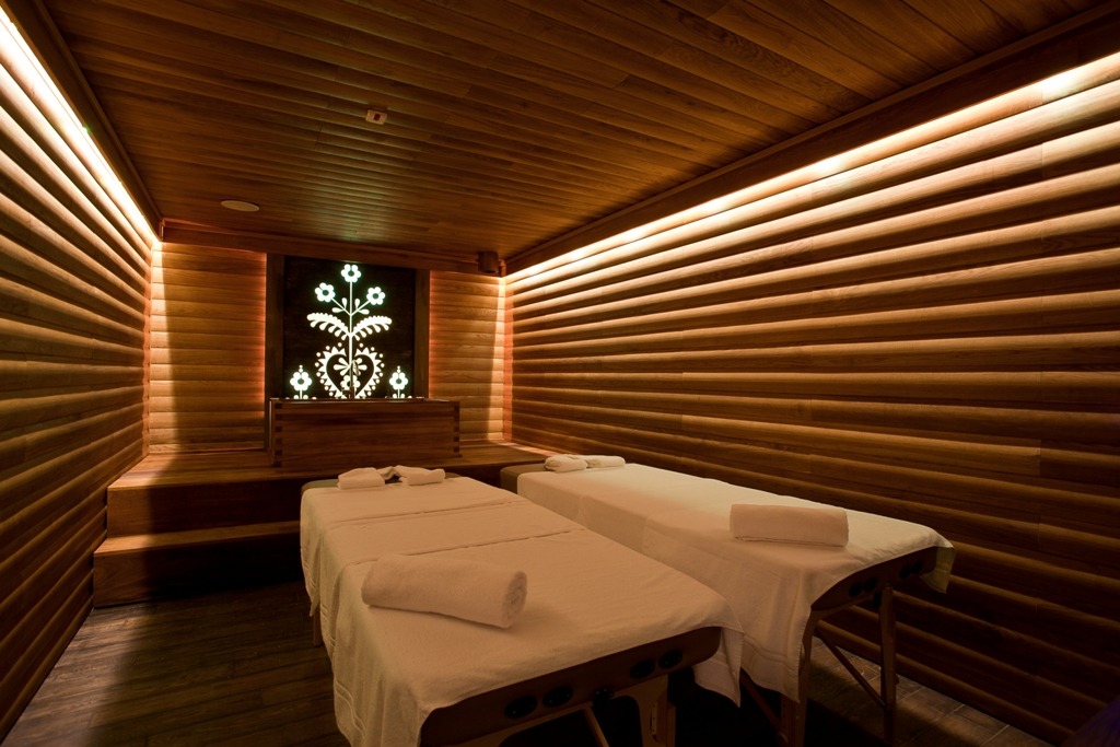 Drewniane wnętrze spa z dwoma przygotowanymi leżankami do masażu, dekoracyjnym podświetlanym panelem z motywem roślinnym i ciepłym oświetleniem.
