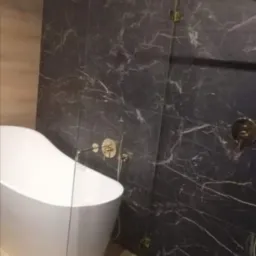 Nowoczesna łazienka z wolnostojącą białą wanną, czarnymi marmurowymi płytkami na ścianach i podłodze, złotymi bateriami i szklanym panelem prysznicowym.