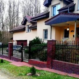 Czarno-metalowe ogrodzenie z dekoracyjnymi elementami, osadzone na niskim murku z czerwonej cegły, otaczające beżowy dom z ciemną dachówką i numerem 15, widoczne drzewa bez liści w tle.