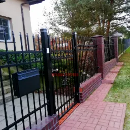 Czarna brama z metalowymi prętami i ozdobnymi elementami, zamontowana na ceglanych słupkach, z czarną skrzynką na listy, prowadząca do posesji z kostki brukowej i trawnika.