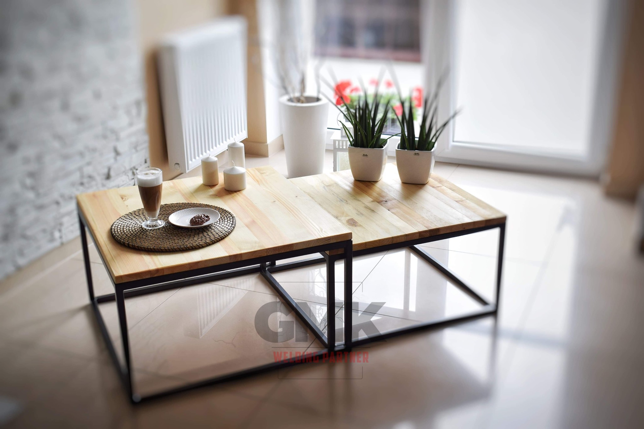 Dwa stoliki kawowe z drewnianymi blatami i czarnymi metalowymi nogami w stylu loft, na jednym z nich szklanka z kawą latte i talerzyk z ciastkiem, w tle okno i rośliny doniczkowe.