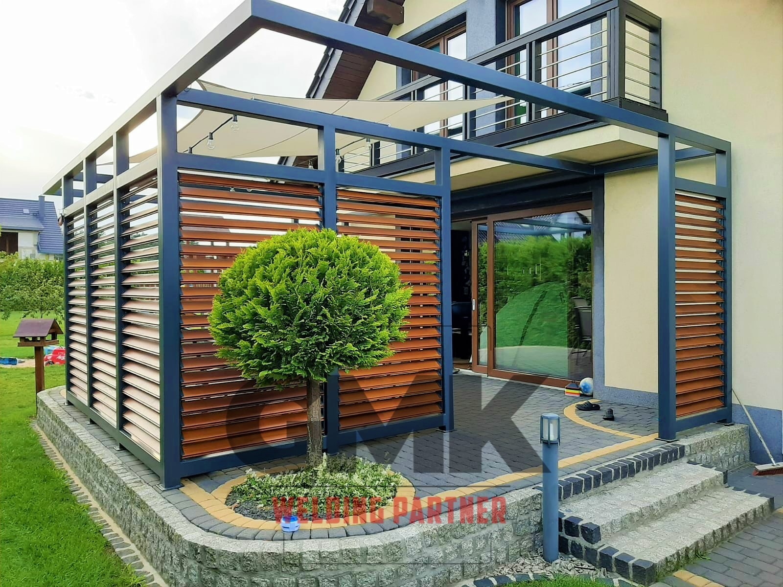 Nowoczesna pergola z metalową ramą w kolorze grafitowym i drewnianymi lamelami, osłaniająca taras z zielonym drzewkiem kulistym u podstawy, oświetlona girlandą żarówek.