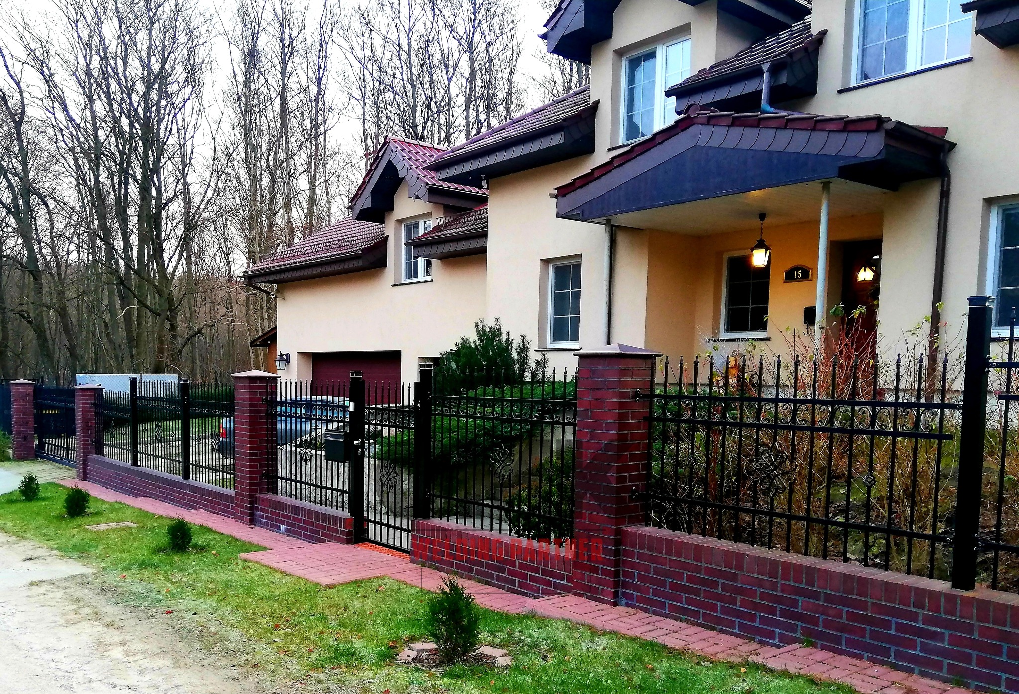 Czarno-metalowe ogrodzenie z dekoracyjnymi elementami, osadzone na niskim murku z czerwonej cegły, otaczające beżowy dom z ciemną dachówką i numerem 15, widoczne drzewa bez liści w tle.