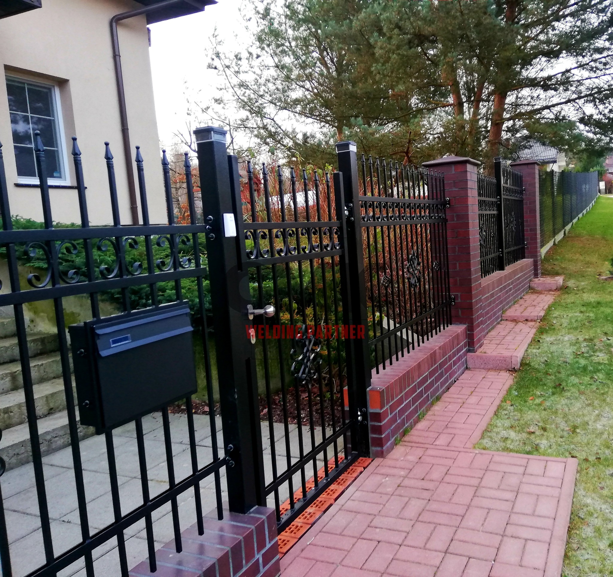 Czarna brama z metalowymi prętami i ozdobnymi elementami, zamontowana na ceglanych słupkach, z czarną skrzynką na listy, prowadząca do posesji z kostki brukowej i trawnika.