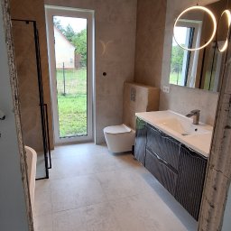 Krzysztof Nawrocki - Jasna łazienka z oknem: beżowe płytki chevron na ścianach, szare płytki podłogowe, toaleta, umywalka z szafką i okrągłe lustro z oświetleniem LED.