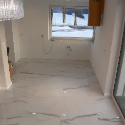 Wnętrze pokoju z nową, białą podłogą z płytek imitujących marmur, widok na okno z zimowym krajobrazem, nowoczesne lampy wiszące i grzejnik na ścianie.