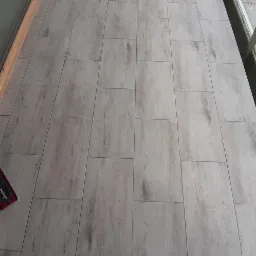 Ułożone panele podłogowe imitujące drewno na balkonie, widok z góry na stopy osoby w roboczych butach, fragment mopa Vileda widoczny w lewym dolnym rogu.