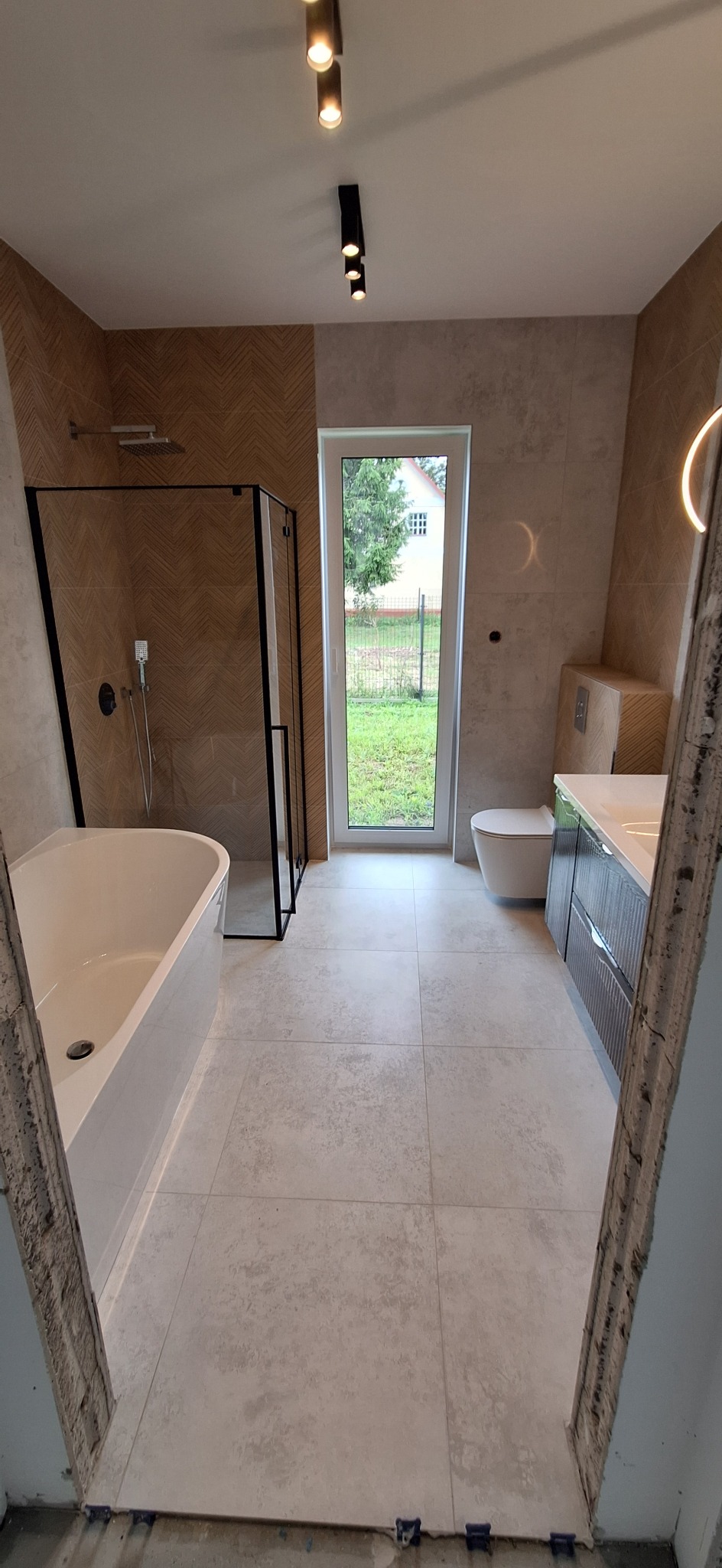 Nowoczesna łazienka z kabiną prysznicową typu walk-in, wanną i umywalką z szafką. Jasne płytki podłogowe i ścienne. Widok na zieleń przez okno.