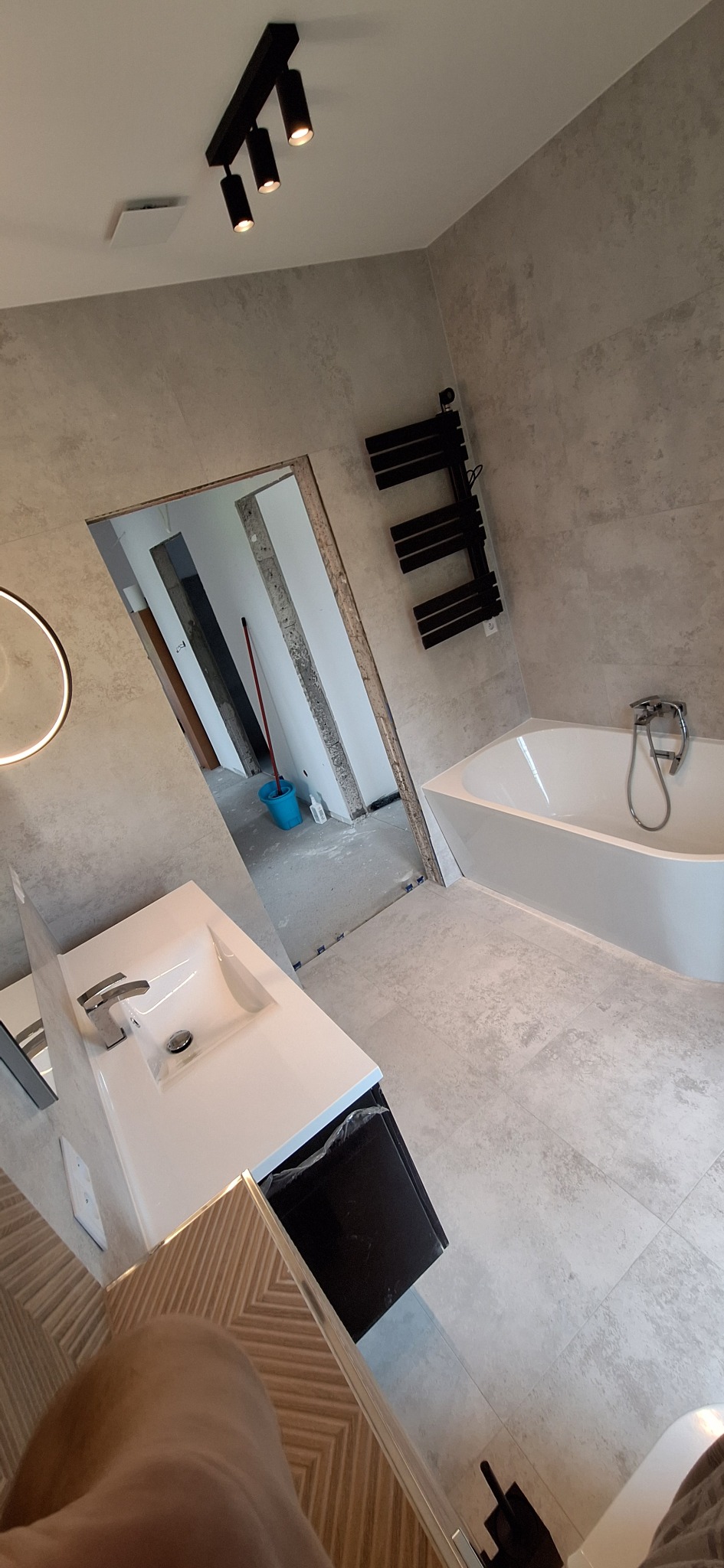Nowoczesna łazienka w trakcie remontu: biała wanna, umywalka z szafką, grzejnik drabinkowy i szare płytki. Widoczny fragment innego pomieszczenia z niebieskim wiadrem.