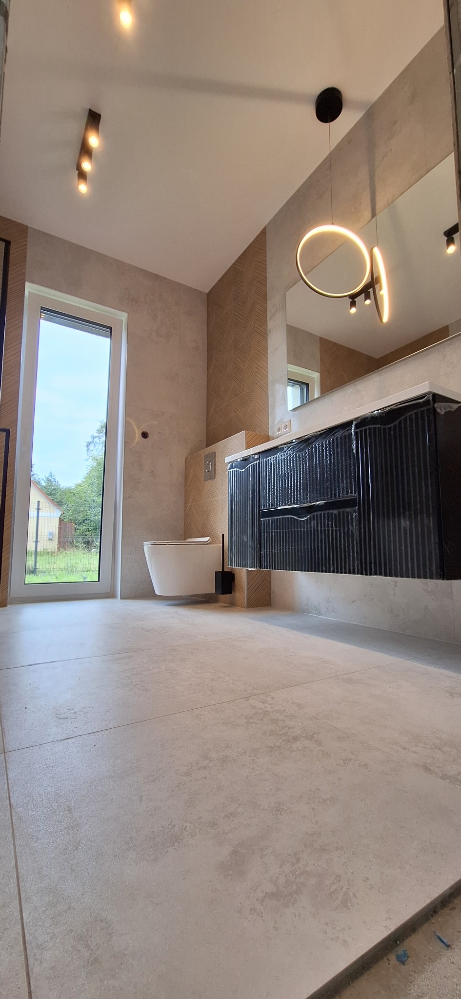 Nowoczesna łazienka z beżowymi płytkami, wiszącą toaletą i czarną szafką pod umywalką. Oświetlenie punktowe i okrągła lampa nad lustrem.