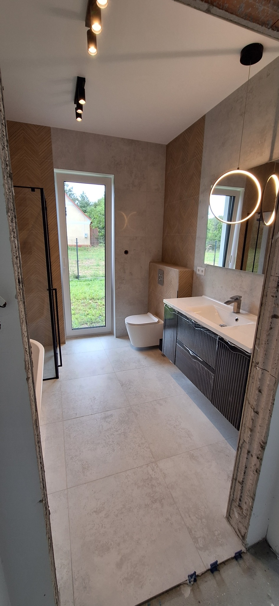 Jasna łazienka z oknem: beżowe płytki chevron na ścianach, szare płytki podłogowe, toaleta, umywalka z szafką i okrągłe lustro z oświetleniem LED.