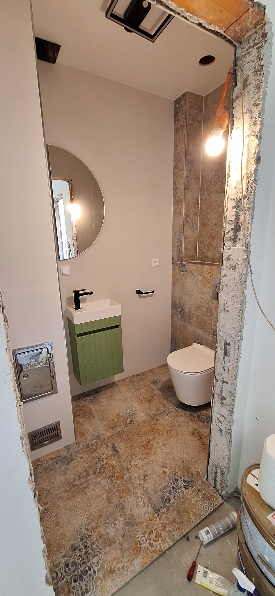 Łazienka w trakcie wykańczania: zielona szafka pod umywalką, okrągłe lustro, płytki podłogowe z wzorem i fragment surowego wejścia z widoczną żarówką.