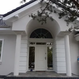 Wejście do domu jednorodzinnego z białymi kolumnami, łukowym nadprożem i szklanymi elementami po bokach drzwi wejściowych, widoczne buty przed drzwiami.