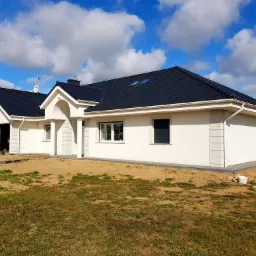 Nowoczesny, parterowy dom z ciemną dach&oacute;wką i białą elewacją, widok z poziomu gruntu na niezagospodarowaną działkę z piaskiem i trawą.