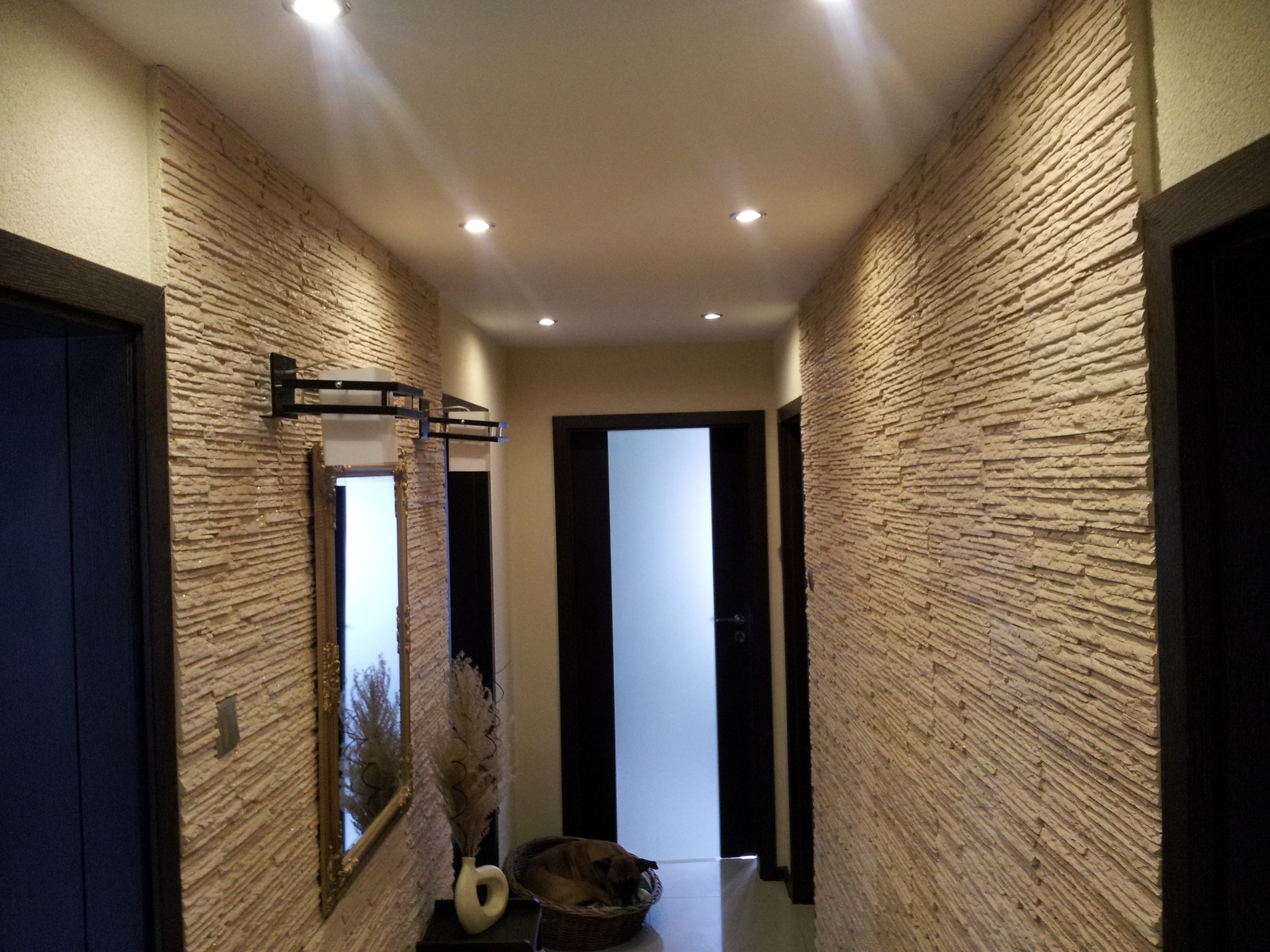 Korytarz z dekoracyjną ścianą z kamienia piaskowego, lustrem z ozdobną ramą, nowoczesnym wazonem z suszem i śpiącym psem w koszyku.