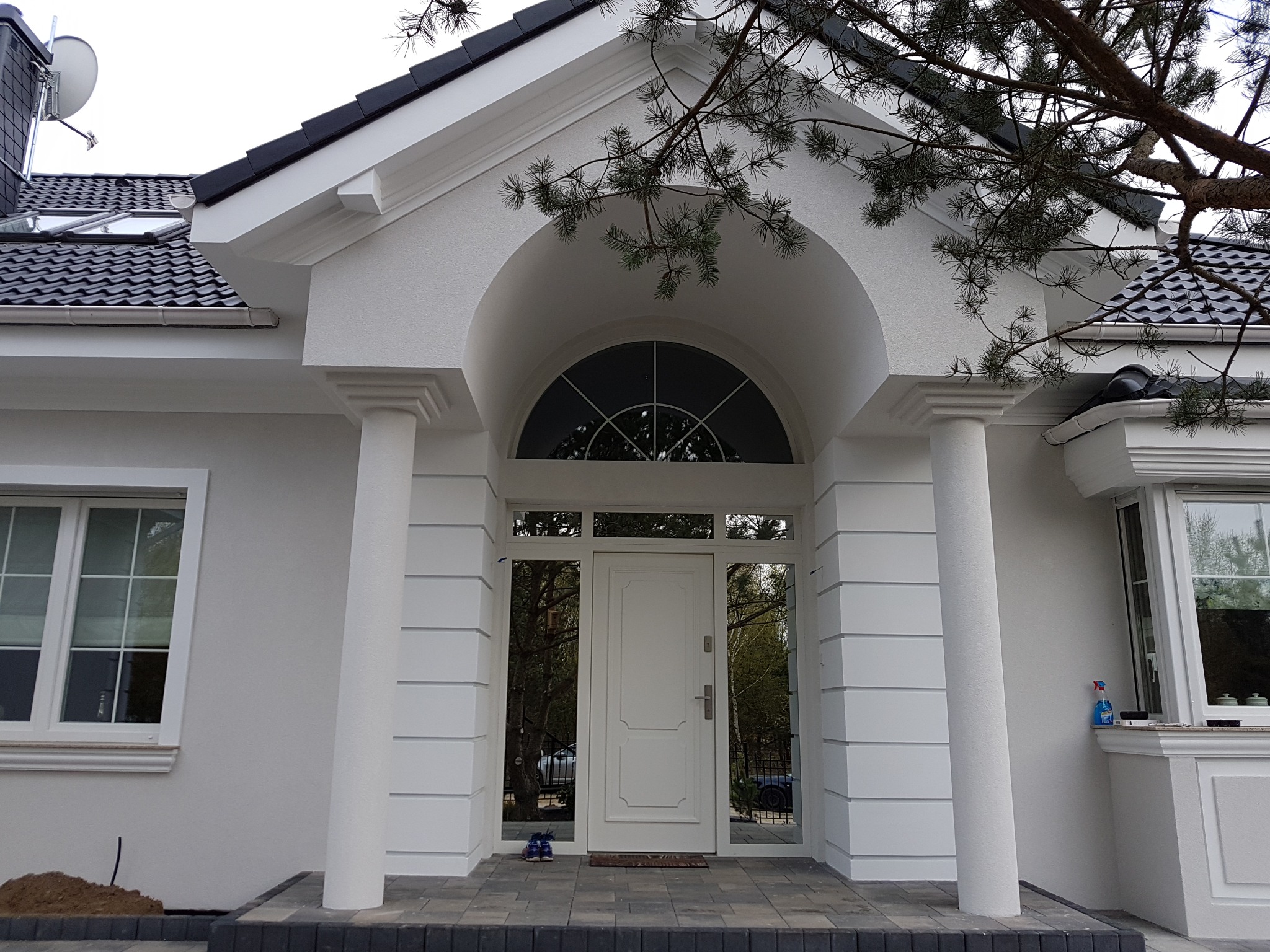 Wejście do domu jednorodzinnego z białymi kolumnami, łukowym nadprożem i szklanymi elementami po bokach drzwi wejściowych, widoczne buty przed drzwiami.