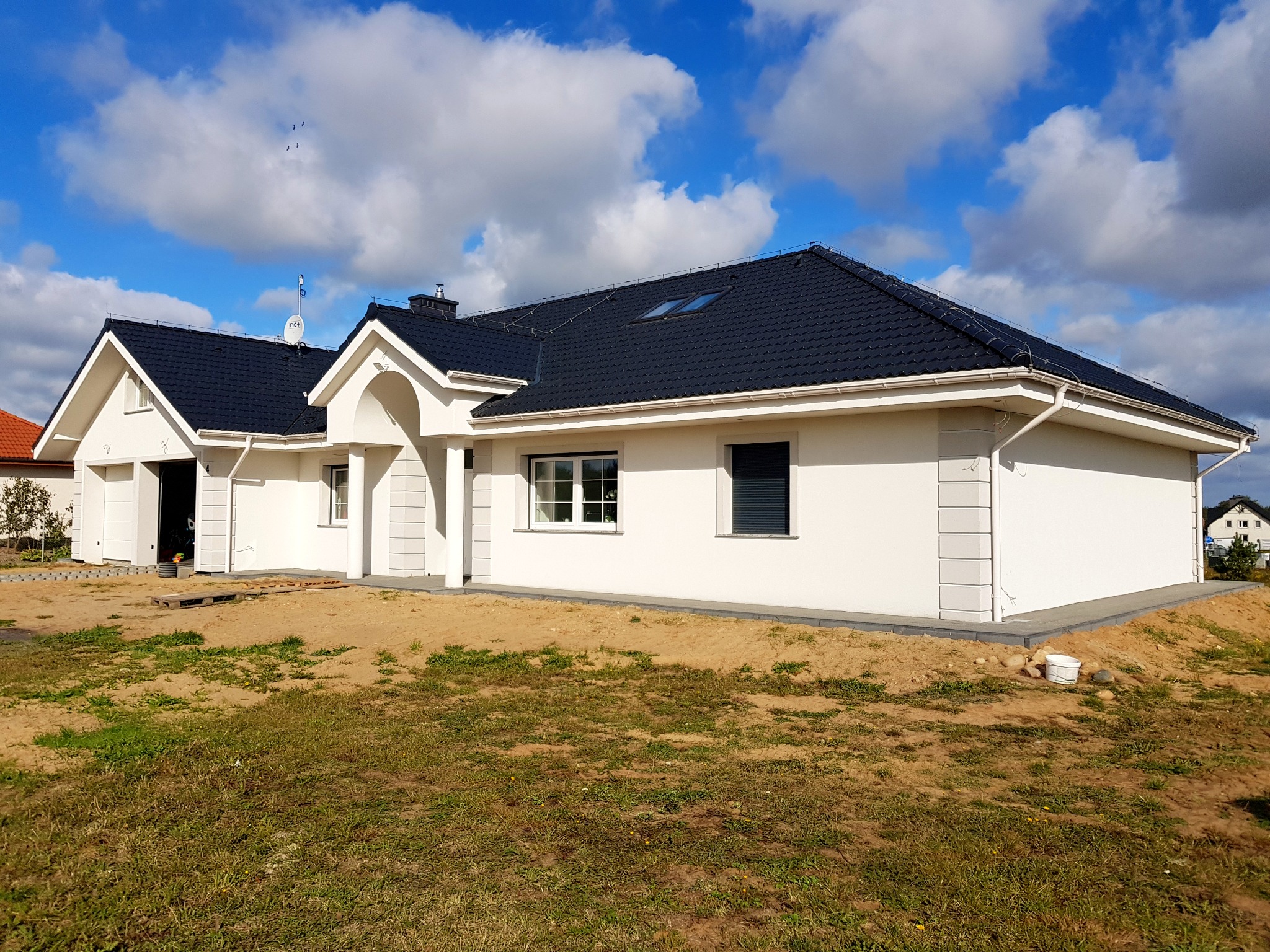 Nowoczesny, parterowy dom z ciemną dachówką i białą elewacją, widok z poziomu gruntu na niezagospodarowaną działkę z piaskiem i trawą.