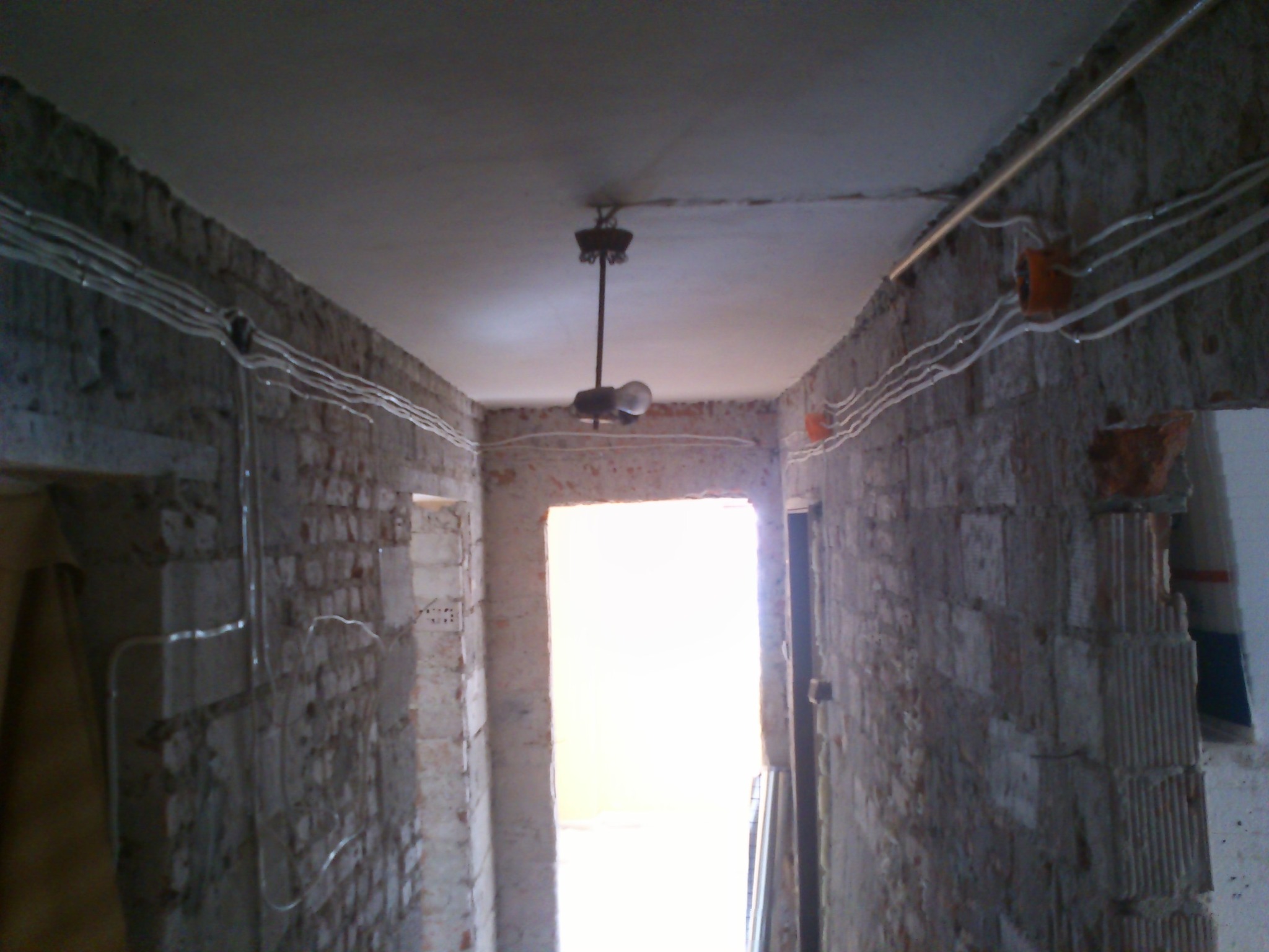 Korytarz w trakcie remontu: surowe ściany z cegły, widoczne przewody elektryczne w listwach, centralnie umieszczona lampa wisząca z żarówką, przejście do jasnego pomieszczenia.