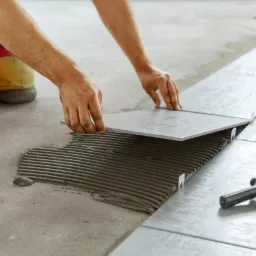 Układanie szarych płytek podłogowych z użyciem krzyżyków dystansowych i narzędzia do poziomowania, widoczna warstwa kleju na podłożu, pracownik w czerwono-żółtych spodniach.