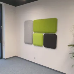 Cztery panele akustyczne w kolorach szarym, zielonym i czarnym zamontowane na białej ścianie w biurze, z widocznym oknem i rośliną doniczkową.
