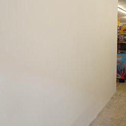 Usługi Ogólnobudowlane Sebastian Ressel - Pomalowana na biało ściana w pomieszczeniu handlowym, widoczne płytki podłogowe i fragment regału z zabawkami w tle.