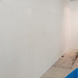 Usługi Ogólnobudowlane Sebastian Ressel - Świeżo pomalowana, biała ściana w biurze z widocznymi śladami po mocowaniach na górze, sufit z odkrytą instalacją i fragment podłogi z płytek oraz pojemnik z materiałami reklamowymi w rogu.