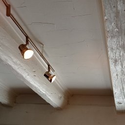 Usługi Ogólnobudowlane Sebastian Ressel - Biały sufit z drewnianymi belkami, oświetlony reflektorami na metalowej szynie, z widoczną fakturą gładzi.