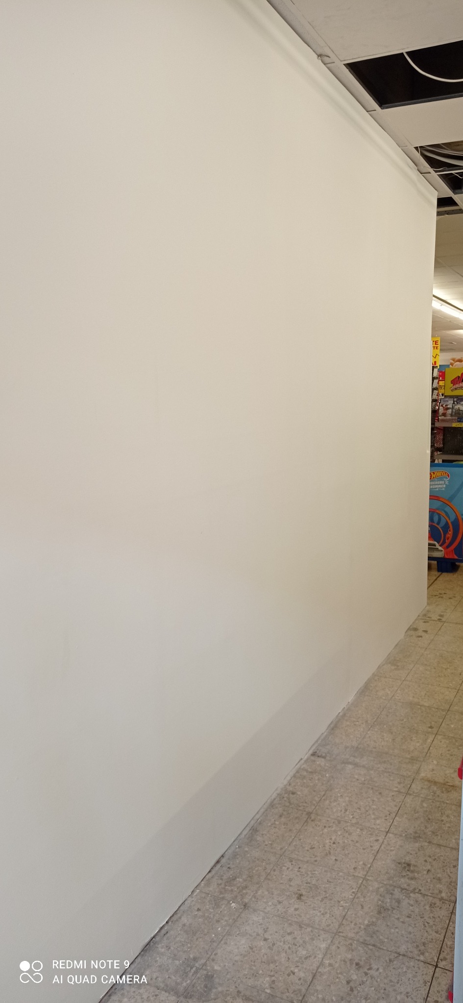 Pomalowana na biało ściana w pomieszczeniu handlowym, widoczne płytki podłogowe i fragment regału z zabawkami w tle.