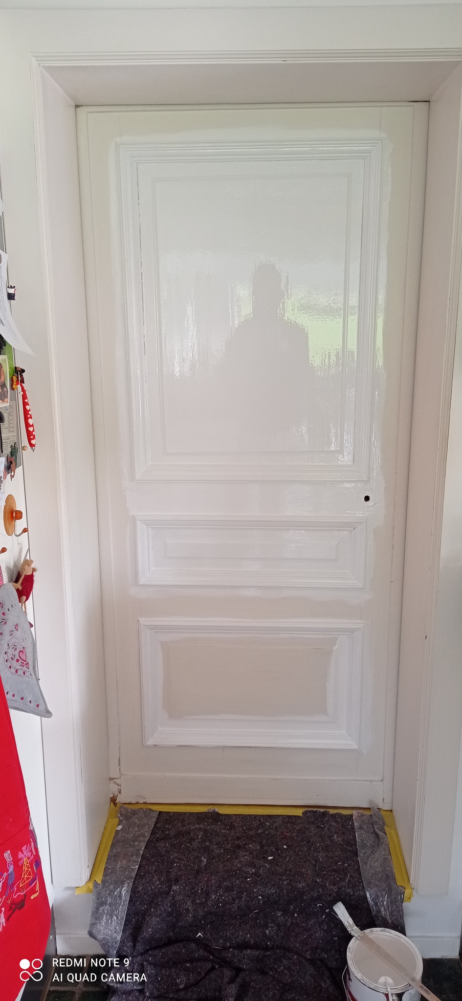 Malowanie białej, drewnianej, panelowej  drzwi wewnętrznych w trakcie renowacji, widoczne ślady świeżej farby, zabezpieczona podłoga i akcesoria malarskie w pobliżu.