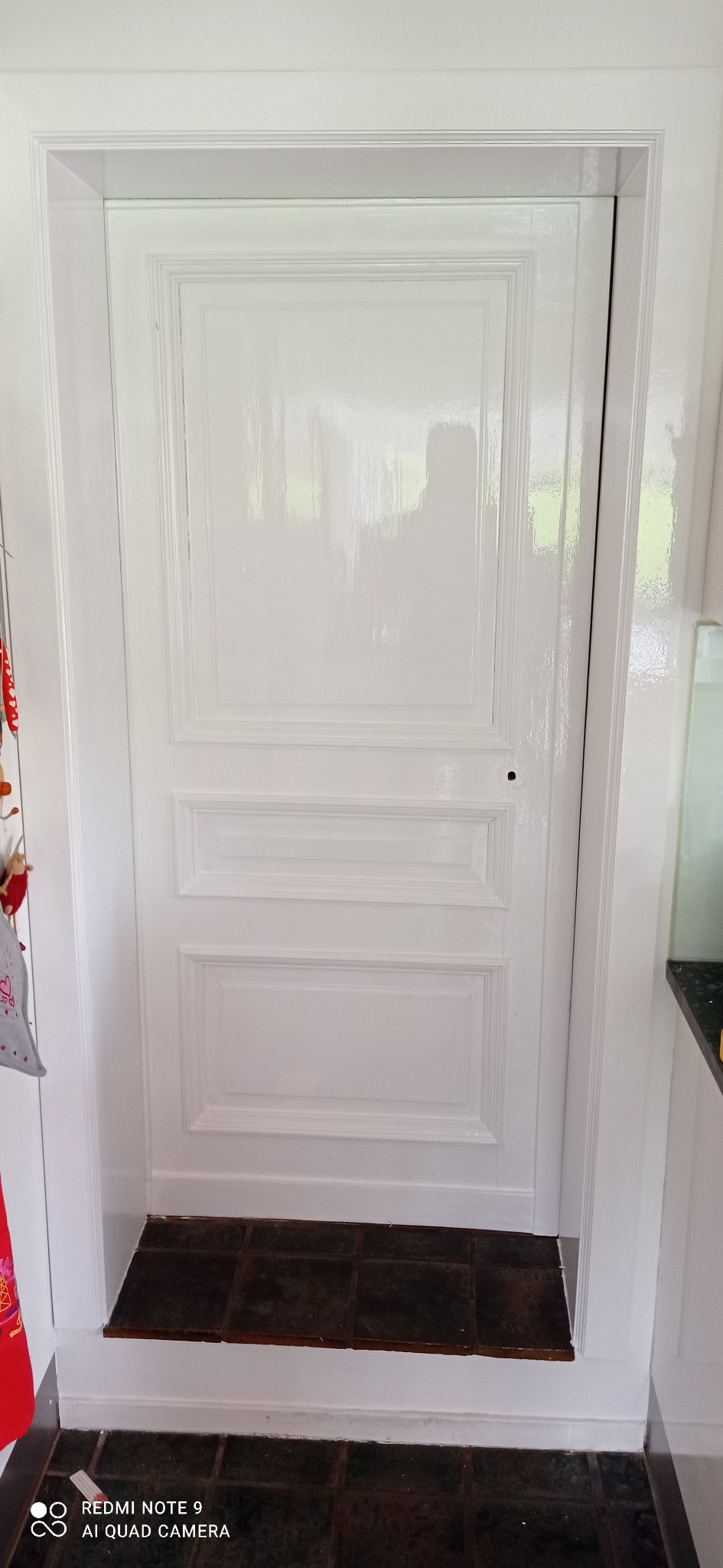 Białe, lakierowane drzwi wewnętrzne z trzema panelami, widoczne od strony pomieszczenia z ciemną, kwadratową podłogą z płytek.