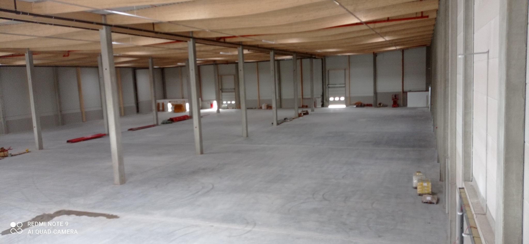 Wnętrze hali magazynowej w stanie surowym, widoczne betonowe słupy, drewniana konstrukcja dachu, ślady po oponach na posadzce, przygotowane materiały budowlane w oddali.