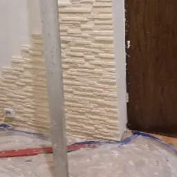 Fragment ściany z panelami imitującymi kamień, w trakcie montażu, zabezpieczona folią malarską, widoczny poziomica i gniazdko elektryczne.