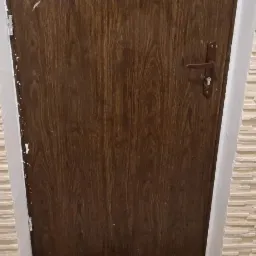 Ciemnobrązowe drzwi z brązową klamką, obok ściana wykończona dekoracyjną, beżową okładziną imitującą kamień.