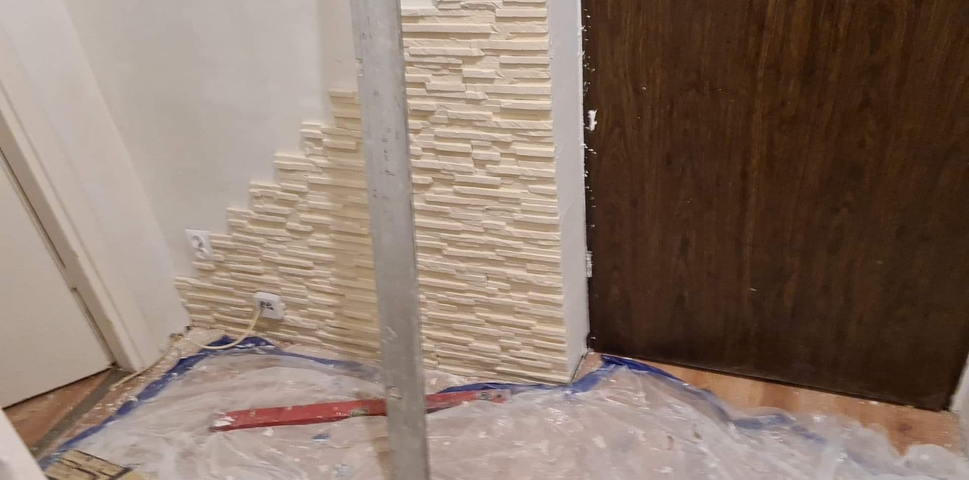 Fragment ściany z panelami imitującymi kamień, w trakcie montażu, zabezpieczona folią malarską, widoczny poziomica i gniazdko elektryczne.