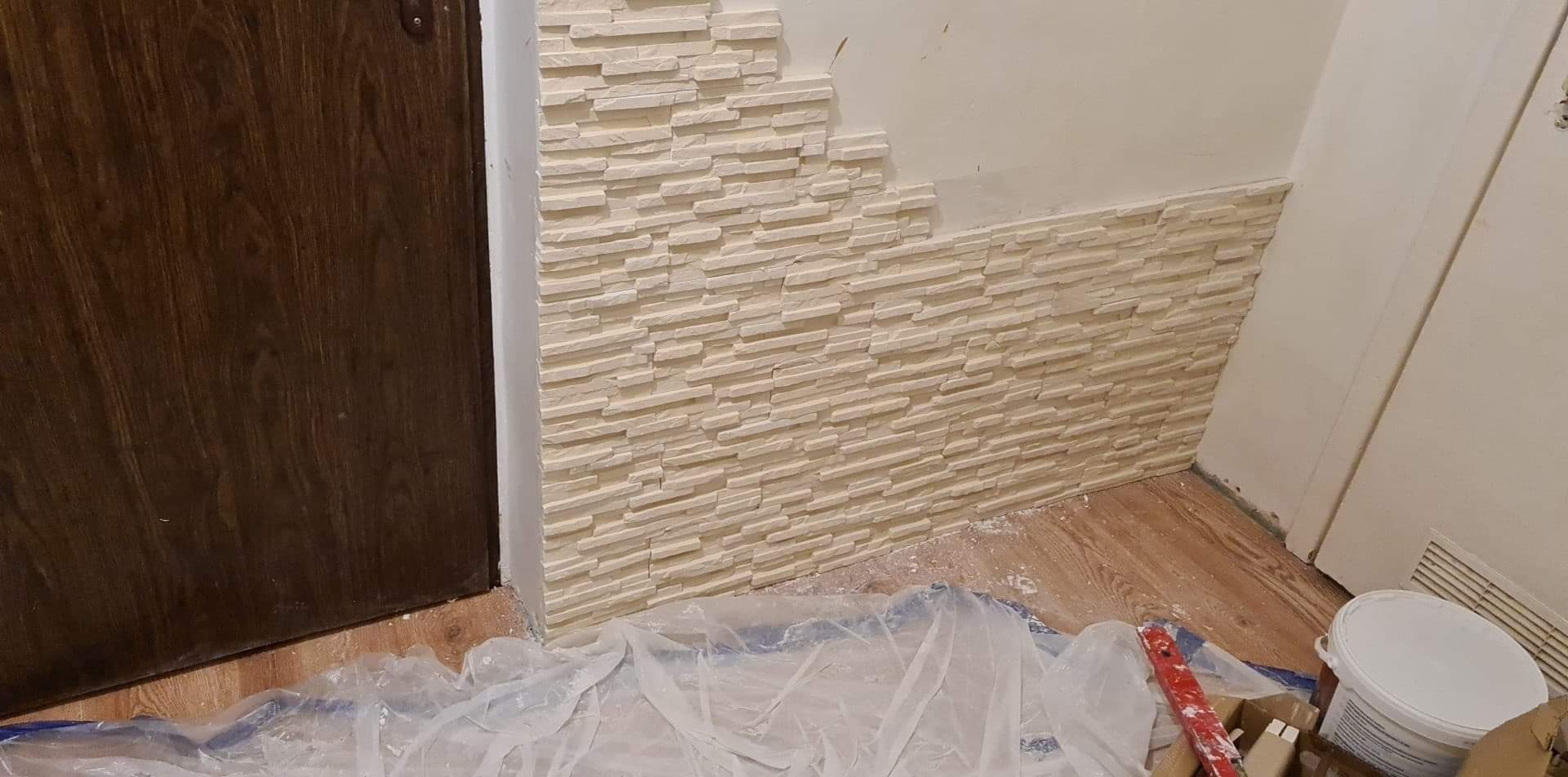 Fragment ściany z częściowo ułożoną dekoracyjną okładziną kamienną w odcieniach beżu, widoczne narzędzia i zabezpieczenia podłoża folią malarską.